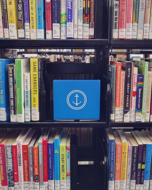 A bookshelf in a library's self-help section displays a bright blue "Find Your Anchor" box with a white anchor symbol in the center. The box stands out among a variety of colorful book spines, including titles like Game Changers, Start More Than You Can Finish, and How to Be Fine.